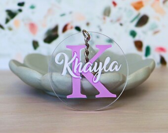 May include: A clear acrylic keyring with a pink letter K and the name "Khayla" written in white. The keyring is on a light wood surface.