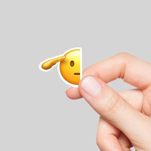 May include: A hand holding a yellow emoji sticker with a single eyebrow raised and a hand over its eye.