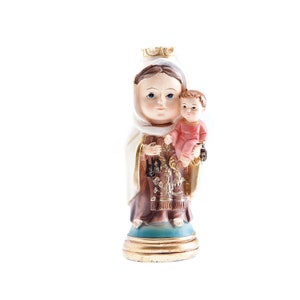 Puede incluir: Una figurita de la Virgen María sosteniendo a un bebé Jesús. La Virgen María lleva una túnica blanca con una faja de color marrón y dorado. El bebé Jesús lleva una túnica rosa. La figurita está hecha de resina y tiene una base dorada.