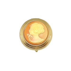 Può includere: Un compatto rotondo color oro con un cammeo in rilievo di un profilo femminile. Il cammeo è in tonalità arancio e bianco.