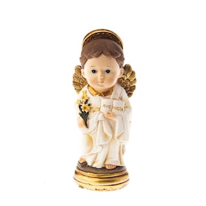 Può includere: Una piccola statuina bianca di un angelo con ali e aureola dorate. L'angelo tiene un libro che dice "Ave Maria" e un mazzo di fiori gialli.