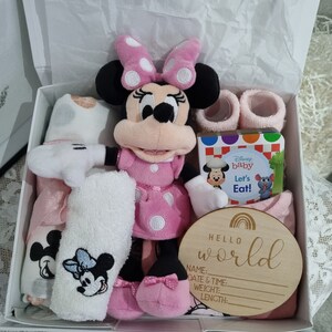 May include: A pink and white polka dot Minnie Mouse plush toy, a pink and white baby blanket, a white towel with a Minnie Mouse embroidery, a pink and white baby outfit, a pink and white baby sock, a book titled "Disney Baby Let's Eat!" and a wooden sign that says "Hello World" with blank lines for name, date and time, weight and length.