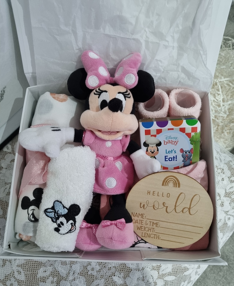 May include: A pink and white polka dot Minnie Mouse plush toy, a pink and white baby blanket, a white towel with a Minnie Mouse embroidery, a pink and white baby outfit, a pink and white baby sock, a book titled "Disney Baby Let's Eat!" and a wooden sign that says "Hello World" with blank lines for name, date and time, weight and length.