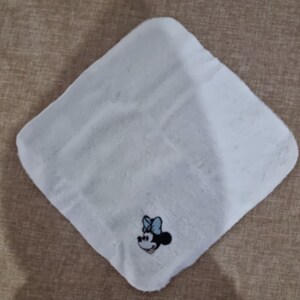 May include: White, soft, square cloth with an embroidered image of Minnie Mouse in the center. Minnie Mouse is wearing a blue bow and has a black outline.