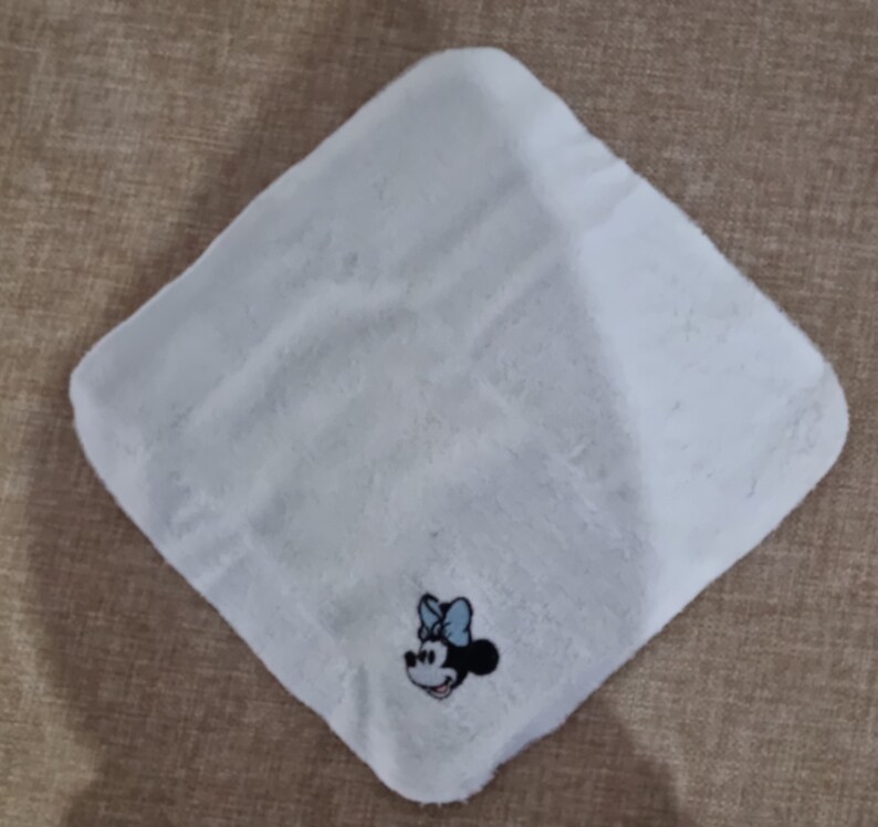 May include: White, soft, square cloth with an embroidered image of Minnie Mouse in the center. Minnie Mouse is wearing a blue bow and has a black outline.