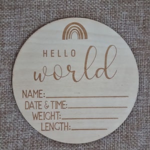 May include: A round wooden plaque with a rainbow design and the text "Hello world". The plaque has blank lines for writing the baby's name, date and time of birth, weight, and length.
