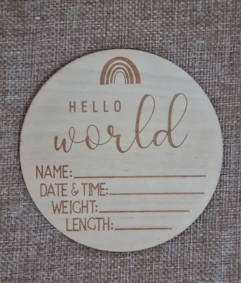 May include: A round wooden plaque with a rainbow design and the text "Hello world". The plaque has blank lines for writing the baby's name, date and time of birth, weight, and length.
