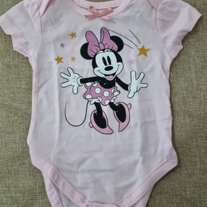 May include: Pink baby onesie with a graphic of Minnie Mouse wearing a pink bow and white polka dot skirt. The onesie has gold stars and a pink bow on the neckline.