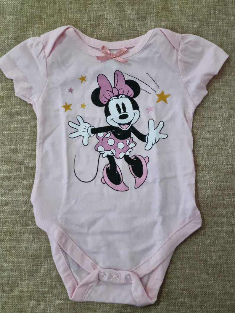 May include: Pink baby onesie with a graphic of Minnie Mouse wearing a pink bow and white polka dot skirt. The onesie has gold stars and a pink bow on the neckline.