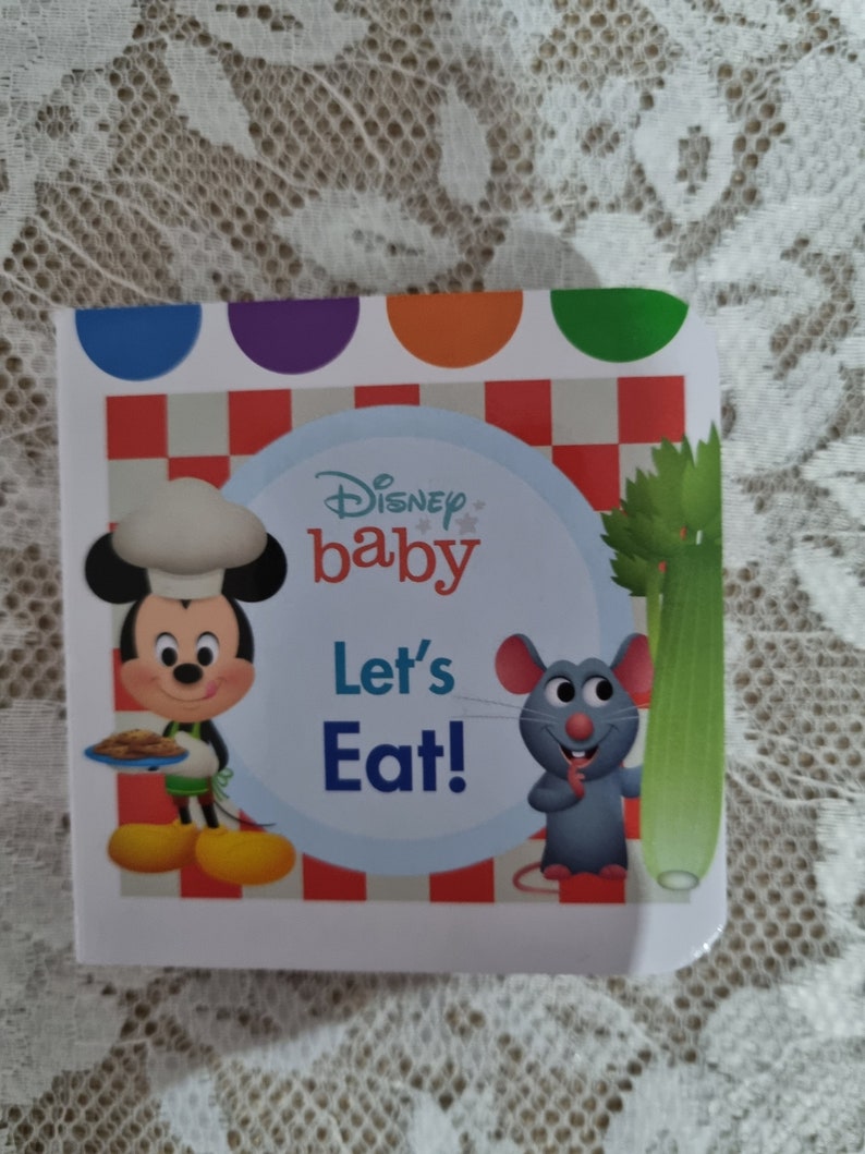 May include: A Disney Baby board book with a red and white checkered background. Mickey Mouse is dressed as a chef and holding a plate of food. A gray mouse is holding a stalk of celery. The book title is "Let's Eat!"