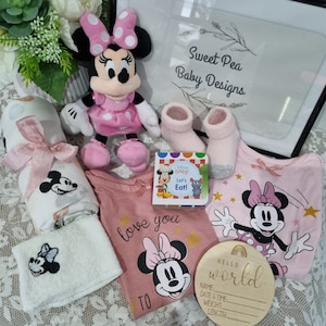 May include: A pink and white baby gift set with a Minnie Mouse theme. The set includes a plush Minnie Mouse toy, a pink and white blanket, a pink and white onesie with a Minnie Mouse graphic, a pair of pink socks, a book, and a wooden sign that says "Hello World".