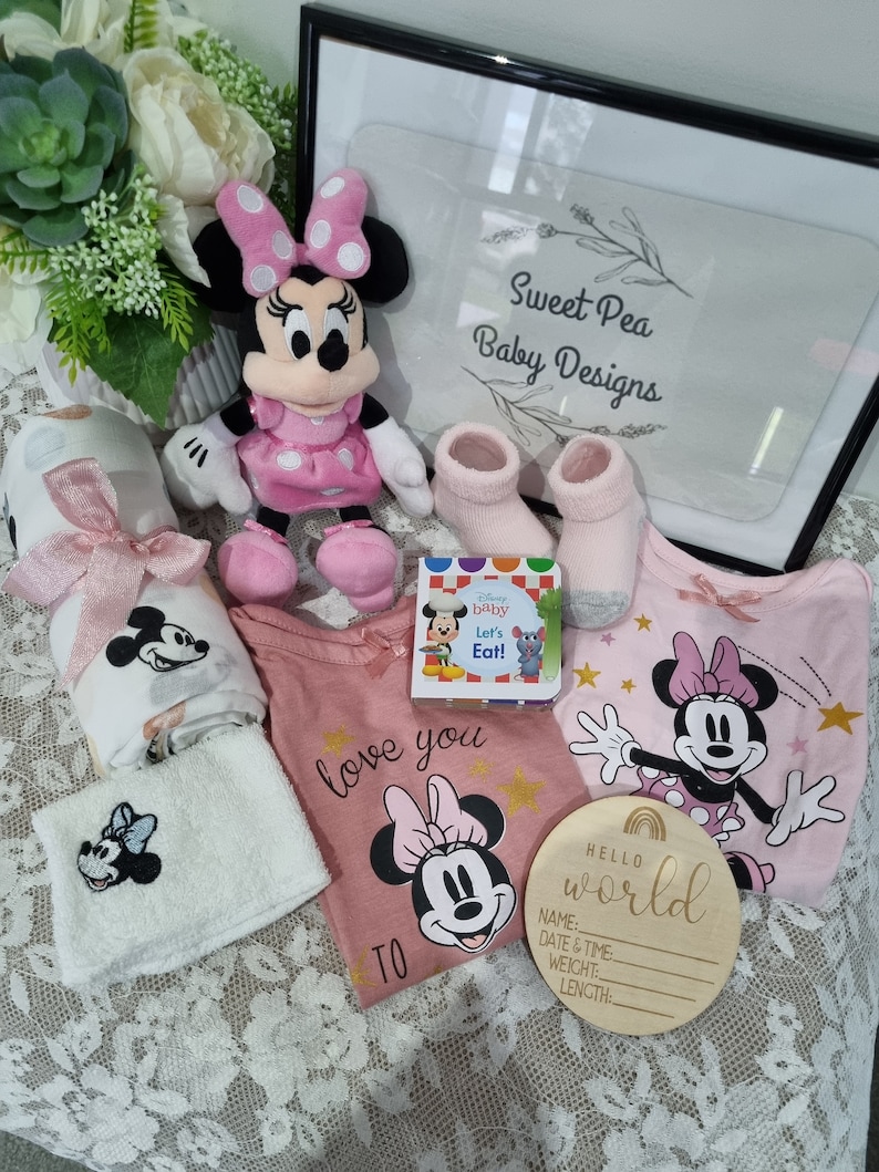 May include: A pink and white baby gift set with a Minnie Mouse theme. The set includes a plush Minnie Mouse toy, a pink and white blanket, a pink and white onesie with a Minnie Mouse graphic, a pair of pink socks, a book, and a wooden sign that says "Hello World".