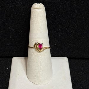 May include: A gold ring with a pink oval-shaped gemstone and a smaller diamond accent. The ring is on a white ring display stand.
