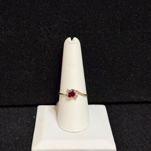 Peut inclure: Une bague en or avec une pierre ovale rouge et deux petites pierres blanches. La bague est sur un présentoir blanc.