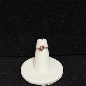 May include: A gold-toned ring with a cluster of small, red gemstones and a few white gemstones. The ring is displayed on a white ring holder against a black background. The gemstones are arranged in a floral pattern.