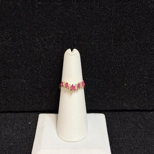 May include: A gold band ring with a curved row of pink gemstones. The ring is on a white display stand.