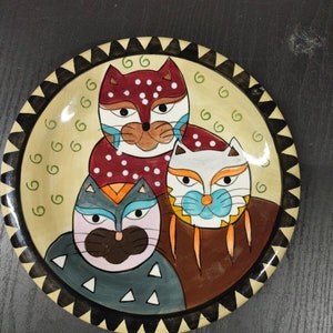 May include: A ceramic plate with a black and white border and a beige background. Three stylized cats are painted on the plate. The cats are red, white, and blue with black eyes and brown noses. The plate is a decorative item.
