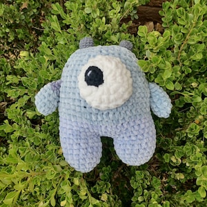May include: A crocheted blue and white plush toy shaped like a monster with one large eye. The toy is sitting on a bed of green leaves.