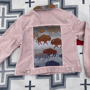 May include: A pink denim jacket with a woven panel on the back featuring a pattern of brown and grey buffalo. The panel is framed with a dark blue border.