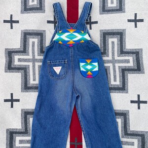Puede incluir: Mono vaquero azul con un diseño geométrico colorido en el peto y el bolsillo trasero. El mono tiene tirantes ajustables y un logotipo de Guess en el bolsillo trasero. El mono se muestra sobre una alfombra estampada.