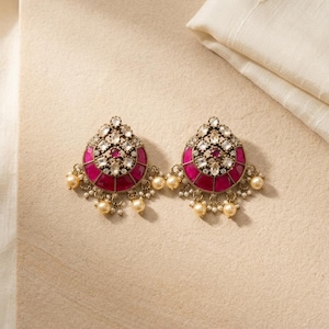 May include: A pair of gold-toned earrings featuring a teardrop shape with deep pink enamel panels, clear faceted stones, and small dangling cream-coloured pearls. The metal has an antique finish with intricate detailing around the central stones.