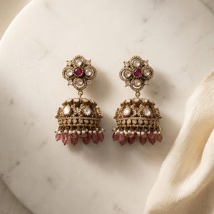 May include: A pair of gold-toned jhumka earrings featuring a floral-shaped stud with a central red stone, surrounded by clear crystals. The bell-shaped drop is adorned with intricate crystal detailing and finished with dangling pink beads.