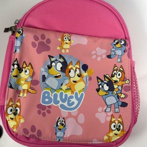 Op de afbeelding: Roze rugzak met de animatiefiguren Bluey en Bingo. De voorzak heeft een roze achtergrond met pootafdrukken en cartoonafbeeldingen van de personages. Het woord "Bluey" is in het blauw op de voorkant gedrukt.