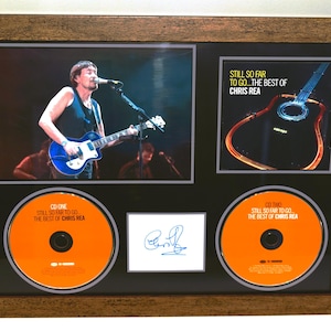 Può includere: Esposizione incorniciata di cimeli di Chris Rea. Include una foto del concerto, l'artwork della copertina dell'album, due CD e un autografo. I CD sono arancioni con testo nero. La cornice è marrone.