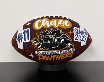 May include: A brown football featuring white laces and custom hand-painted designs including the name Chase, the number 11, the text Senior 2027, and Southwest Dekalb Panthers with a black panther graphic, accented by small white and blue stars.