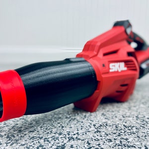 May include: A red and black cordless leaf blower with a black nozzle and a red tip. The blower has the word "SKIL" printed on the side.