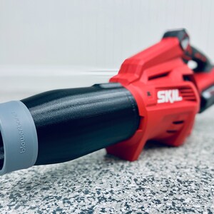 May include: A red and black cordless leaf blower with a gray nozzle. The nozzle has the word "Blowforce" printed on it. The leaf blower has the word "SKIL" printed on the side.
