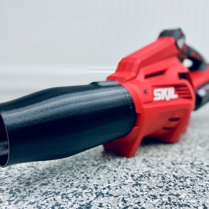 May include: A red and black cordless leaf blower with a black nozzle. The blower has the word "SKIL" printed on the side.