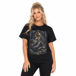 Può includere: Una t-shirt nera a maniche corte con una stampa grafica di una figura in abito scuro che scende una scala di pietra sotto un grande orologio. Il design include una cornice gotica decorata e il testo THE MIDNIGHT SPELL nella parte inferiore.