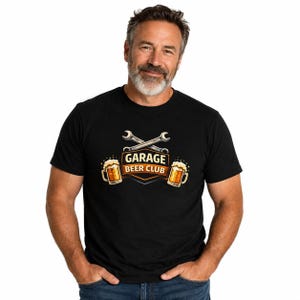 May include: A black short-sleeved t-shirt featuring a graphic print of two crossed silver wrenches above a shield emblem with the text GARAGE BEER CLUB in bold letters, flanked by two frothy mugs of golden beer on either side of the central design.