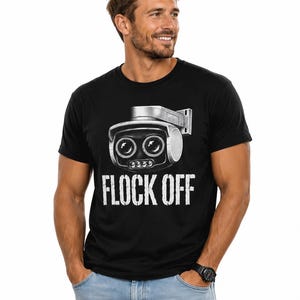 May include: A black short-sleeved t-shirt featuring a graphic of a silver security camera with two large lenses and the text "FLOCK OFF" printed in white capital letters underneath the camera illustration on the front of the garment.
