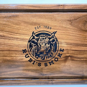 May include: A wooden serving tray with black metal handles. The tray has a dark wood grain and is engraved with the text "EST. 1989 RUBI'S SMOKE" and a bull's head logo.