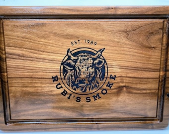 May include: A wooden serving tray with black metal handles. The tray has a dark wood grain and is engraved with the text "EST. 1989 RUBI'S SMOKE" and a bull's head logo.