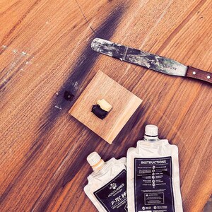 May include: Two tubes of epoxy resin with instructions on one tube. The tubes are on a wooden surface with a small piece of epoxy resin on a separate piece of wood. A metal putty knife is also on the surface.