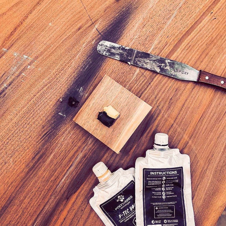 May include: Two tubes of epoxy resin with instructions on one tube. The tubes are on a wooden surface with a small piece of epoxy resin on a separate piece of wood. A metal putty knife is also on the surface.
