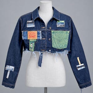 May include: A dark blue denim cropped jacket featuring a frayed hem, a light green chest pocket, and various rectangular fabric patches in shades of blue and white. A tan Levi Strauss & Co. brand label is stitched onto the front left side.