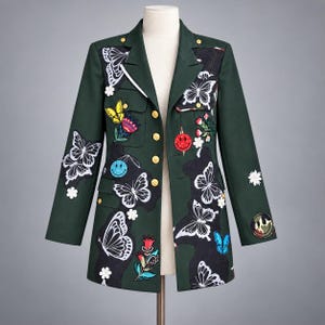 May include: A dark green blazer featuring a notched lapel, gold-tone buttons, and various embroidered patches including white butterflies, colorful flowers, and smiley faces. The jacket is displayed on a mannequin and has a structured silhouette.