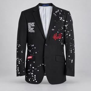 May include: A black blazer featuring a white paint splatter pattern, a two-button closure, and a light grey interior lining. The front includes a rectangular patch with text and a red embroidered script word Hustle, plus a small red Original patch.