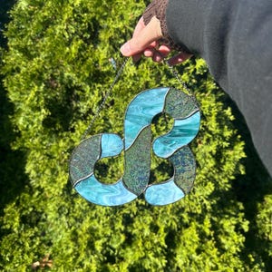 May include: A stained glass art piece in the shape of the letter "S". The glass features shades of teal and green with a swirling pattern. The piece is held by a silver chain against a backdrop of green foliage.