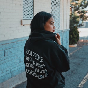 K&ouml;nnte beinhalten: Ein schwarzer Kapuzenpullover mit den Worten "GOOD PEOPLE, GOOD NIGHTS, GOOD HIGHS, GOOD HEALTH" in Wei&szlig; aufgedruckt. Die Person tr&auml;gt schwarze Shorts und steht drau&szlig;en in der N&auml;he eines Geb&auml;udes mit wei&szlig;en und blauen Akzenten.