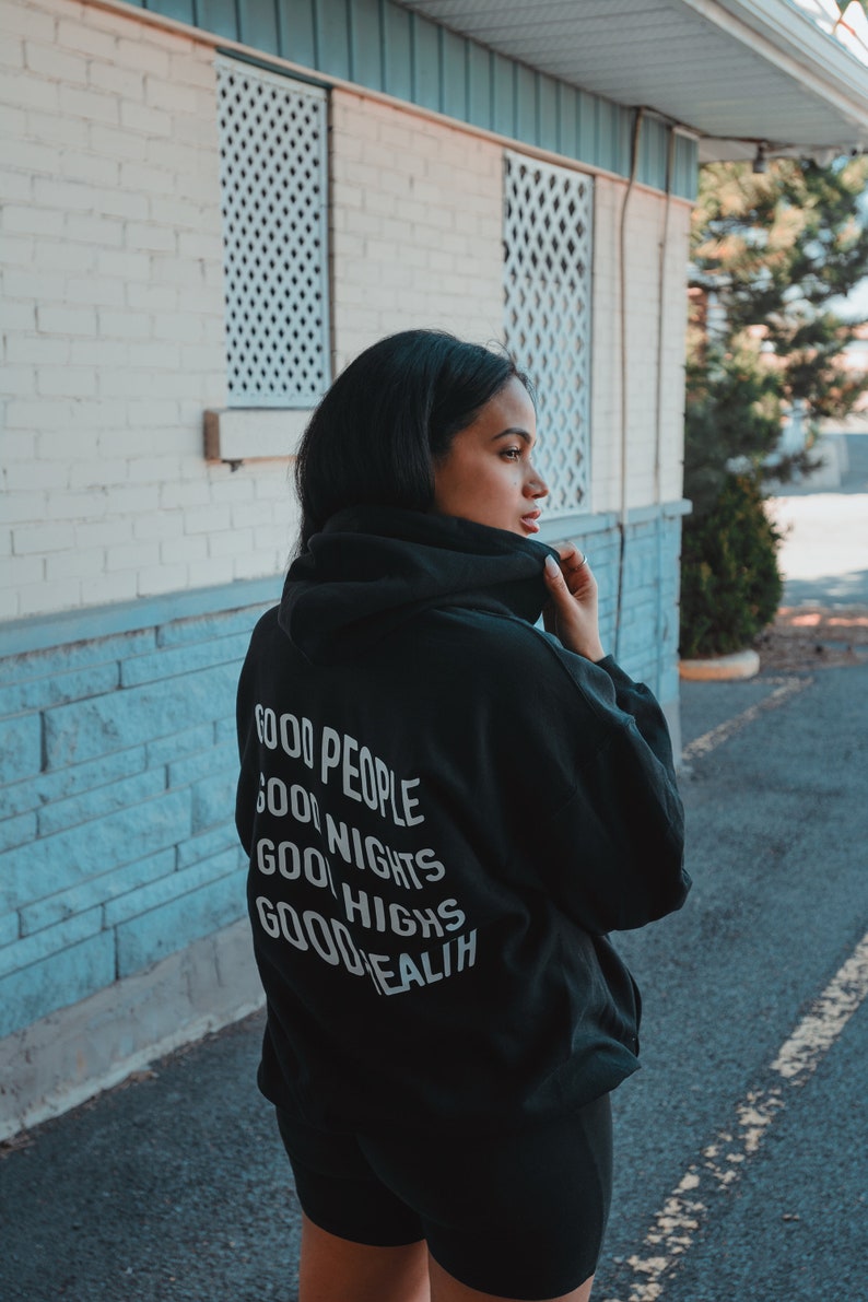 K&ouml;nnte beinhalten: Ein schwarzer Kapuzenpullover mit den Worten "GOOD PEOPLE, GOOD NIGHTS, GOOD HIGHS, GOOD HEALTH" in Wei&szlig; aufgedruckt. Die Person tr&auml;gt schwarze Shorts und steht drau&szlig;en in der N&auml;he eines Geb&auml;udes mit wei&szlig;en und blauen Akzenten.
