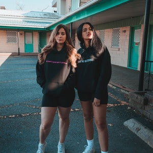 K&ouml;nnte beinhalten: Zwei Personen in schwarzen Kapuzenpullovern und Shorts stehen vor einem Geb&auml;ude. Ein Kapuzenpullover hat den Text "All I want is good music & great sex" in Pink. Der andere Kapuzenpullover sagt "COUNT MY BLESSINGS" in Wei&szlig;.