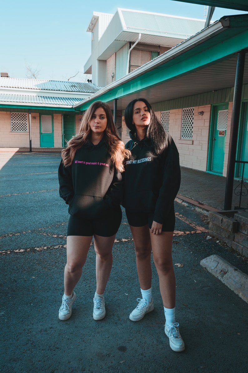 K&ouml;nnte beinhalten: Zwei Personen in schwarzen Kapuzenpullovern und Shorts stehen vor einem Geb&auml;ude. Ein Kapuzenpullover hat den Text "All I want is good music & great sex" in Pink. Der andere Kapuzenpullover sagt "COUNT MY BLESSINGS" in Wei&szlig;.