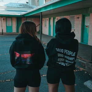 K&ouml;nnte beinhalten: Zwei Personen tragen schwarze Kapuzenpullover und Shorts im Freien. Ein Kapuzenpullover hat eine farbenfrohe Grafik auf dem R&uuml;cken, der andere die wei&szlig;en Worte &bdquo;GOOD PEOPLE GOOD NIGHTS GOOD HIGH&ldquo;. Die Kulisse ist ein Motel.