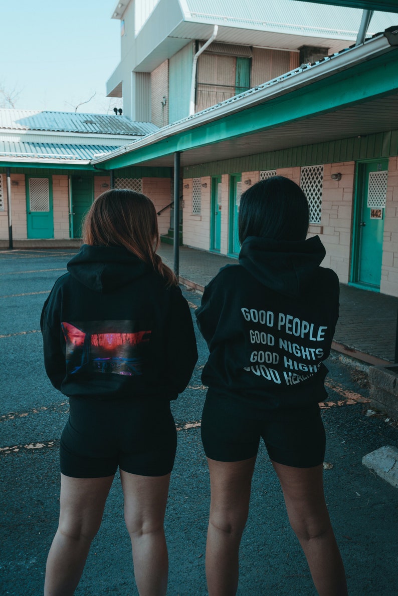 K&ouml;nnte beinhalten: Zwei Personen tragen schwarze Kapuzenpullover und Shorts im Freien. Ein Kapuzenpullover hat eine farbenfrohe Grafik auf dem R&uuml;cken, der andere die wei&szlig;en Worte &bdquo;GOOD PEOPLE GOOD NIGHTS GOOD HIGH&ldquo;. Die Kulisse ist ein Motel.