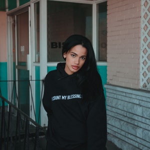 K&ouml;nnte beinhalten: Schwarzer Kapuzenpullover mit dem wei&szlig;en Text "I COUNT MY BLESSINGS". Das Model tr&auml;gt schwarze Shorts. Der Hintergrund zeigt ein Geb&auml;ude mit einer t&uuml;rkisfarbenen und rosa Fassade.